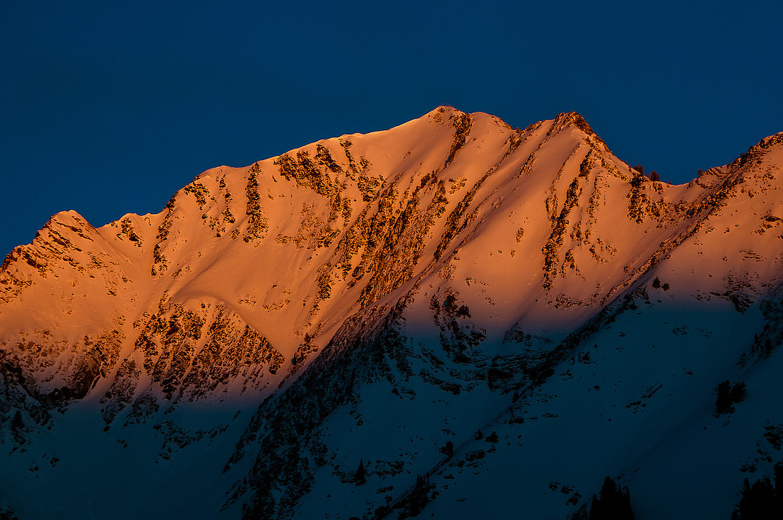 A stunning snow-covered mountain at sunrise, with the top two-thirds illuminated by the morning light and the bottom portion casting a captivating shadow. The bluebird sky and pink hues at the shadow line add to the scenic beauty.