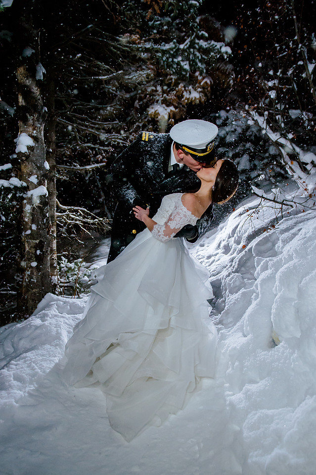 A navy officer in dress blues and white hat is kissing his bride, who wears a wedding dress with delicate lace sleeves and bare shoulders. They stand in falling snow, with snow-covered trees in the background.