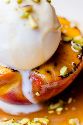 A close-up photo of a barbequed peach with caramelized grill marks on top of a white plate, topped with a scoop of melting vanilla ice cream and sprinkled with crushed pistachios.