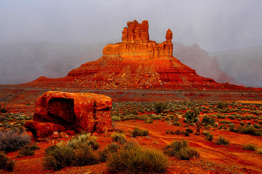 A stunning red rock formation resembling a castle or monastery stands tall amidst the red desert sands of Valley of the Gods, Utah. A smaller red rock is visible in the foreground, and a snowstorm approaches from the background, enhancing the vibrant colors of the scene.