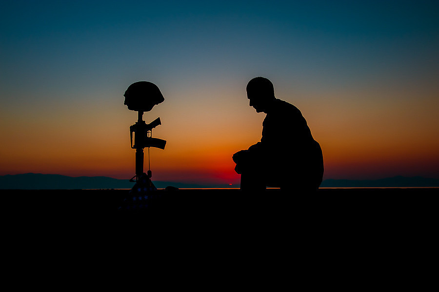 Silhouette of a soldier kneeling at a field cross at sunset.